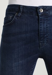 Mörkblå denimjeans med rak passform, femfickorsdesign och silverfärgade detaljer. Tyget har en slät yta med liten blekning.