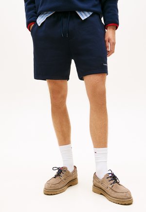 Mann iført marineblå shorts, hvite crew-sokker, brune semskede sko og marineblå genser med synlig stripete skjortekant.