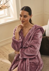 Accappatoio rosa chiaro in peluche con cappuccio e cintura, dotato di maniche lunghe e tasche frontali. Texture morbida e aspetto caldo.