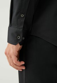 Čierna košeľa s dlhým rukávom, vyrobená z textúrovanej látky, s gombíkovými manžetami s kovovými akcentmi a elegantným, prispôsobeným strihom.