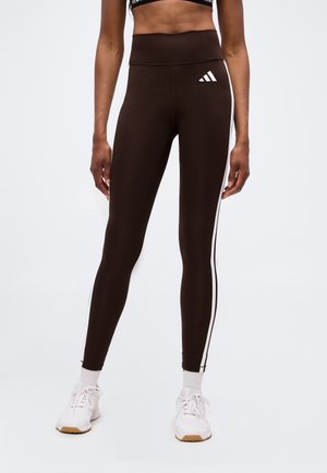 Donna che indossa leggings neri a vita alta con strisce laterali bianche e logo Adidas, abbinati a scarpe sportive bianche e calzini.