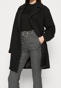 Cappotto di lana nera con cuciture decorative bianche, colletto a contrasto e due tasche frontali, indossato sopra una maglia nera e pantaloni houndstooth.