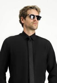 Camisa negra de botones con cuello, que presenta un bolsillo; combinada con una corbata negra con un patrón y gafas de sol negras, textura de tela suave.