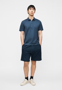 Πόλο μπλε ναυτικού με λεπτομέρειες κουμπιών, συνδυασμένο με σορτς ναυτικού χρώματος. Ελαφρύ ύφασμα, casual σχεδίαση και λευκά αθλητικά παπούτσια. Απλός, μινιμαλιστικός στιλ.