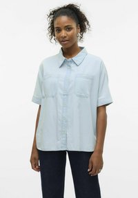 Camicia azzurra chiara a maniche corte in morbido tessuto con bottone frontale, due tasche sul petto e collo. Indossata con jeans scuri.
