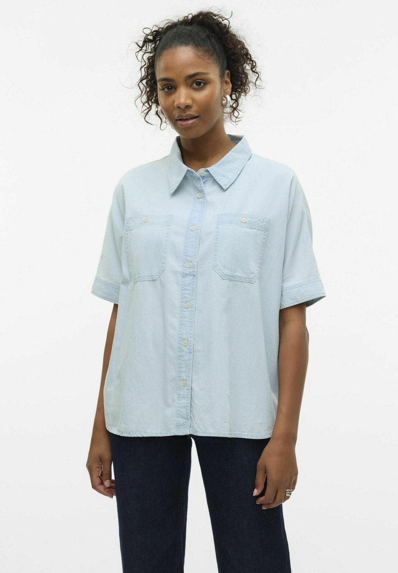 Camicia azzurra chiara a maniche corte in morbido tessuto con bottone frontale, due tasche sul petto e collo. Indossata con jeans scuri.