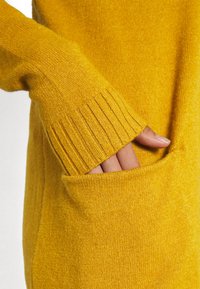 Suéter de manga larga en tejido de punto amarillo brillante, con puños acanalados y un bolsillo frontal donde se ve parcialmente una mano dentro. Textura suave.