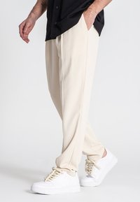Beige, løstsiddende bukser med sidelommer, båret med hvide sneakers. Stofletsningen fremstår glat og let struktureret.