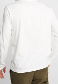 Witte lange mouw shirt gemaakt van katoen. Heeft een ronde hals en een relaxte pasvorm. De stof lijkt zacht met een minimale textuur. Corduroy broeken zichtbaar eronder.