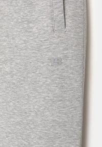 Lys grå stof med et lille præget "PB" logo nær en lodret sidelomme på et beklædningsstykke, der viser en blød tekstur og sømdetaljer.