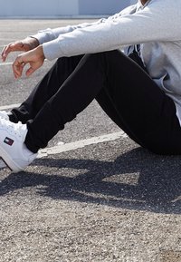 Svart tapered byxor och en ljusgrå hoodie, ihop med vita sneakers. Materialet verkar mjukt; sneakersen har en logotyp på sidan.