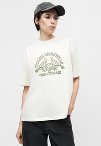 Νεαρό άτομο που φοράει λευκό μπλουζάκι με το λογότυπο "Dickies Workwear Quality Made" και σχέδιο πέταλου αλόγου, καθώς και σκούρο γκρι καπέλο μπέιζμπολ, στέκεται με το ένα χέρι στην τσέπη.