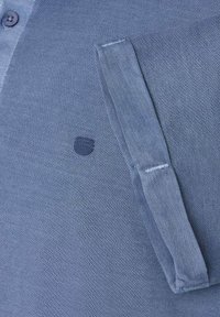 Niebieska polo z fakturowanego materiału z zapięciem na guziki przy kołnierzyku oraz kieszonką naszywaną z logo. Materiał wydaje się oddychający i tkany.