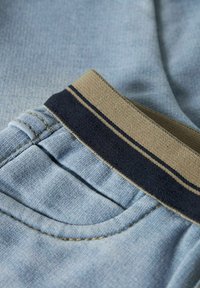 Tela de mezclilla azul claro con una superficie texturizada. La cinturilla es color beige con rayas marinas. Detalles de costura visibles en los bolsillos y las costuras.