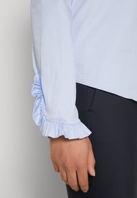 Ljusblå blus med en volangdetalj vid ärmsluten. Tyget ser slätt och lätt ut, kombinerat med mörka, tighta byxor.