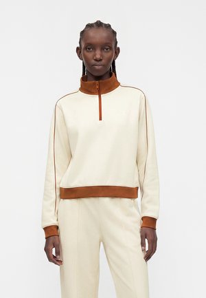 Giovane donna con capelli intrecciati che indossa una felpa beige e marrone con zip a quarto e pantaloni beige abbinati, in piedi davanti a uno sfondo semplice.