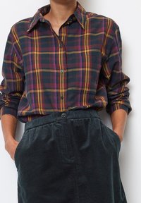 Camicia di cotone a quadri in blu navy, bordeaux e giallo, con colletto a bottone e maniche lunghe. Abbinata a una gonna in velluto a coste verde scuro con texture.