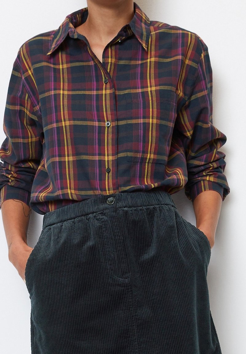 Camicia di cotone a quadri in blu navy, bordeaux e giallo, con colletto a bottone e maniche lunghe. Abbinata a una gonna in velluto a coste verde scuro con texture.
