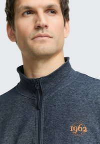 Donkergrijze jas met rits, gemaakt van gestructureerde stof, met een hoge kraag en een oranje cirkelvormig logo met "1962" op het linkerborstgebied.