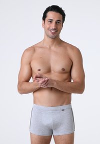 Uomo sorridente senza maglietta in piedi contro uno sfondo bianco, indossando boxer grigi con le mani congiunte davanti.