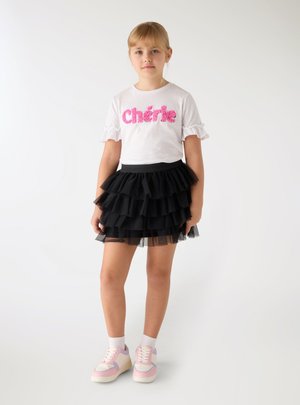 Ragazza giovane in piedi, indossa una maglietta bianca con la scritta rosa "Chérie", gonna in tulle nera a più strati, calzini bianchi e scarpe da ginnastica rosa-bianco.