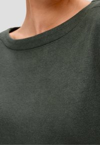 Maglione abbondante verde scuro con scollatura rotonda. Il tessuto sembra morbido con una superficie leggermente testurizzata. Nessun motivo o accento visibile.