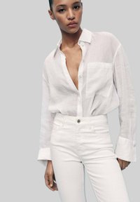 Camicia in lino bianca, dal taglio rilassato, con una tasca sul petto, bottoni a metà e maniche arrotolate, abbinata a jeans bianchi a vita alta.