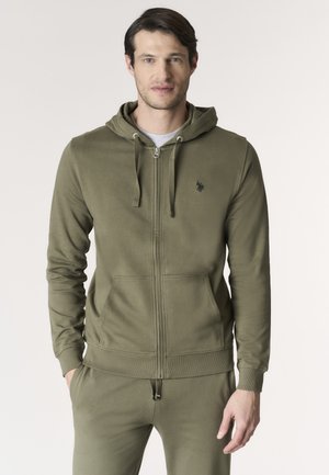 Uomo che indossa una felpa verde oliva con zip e tasche a marsupio e pantaloni coordinati, in piedi su sfondo chiaro.