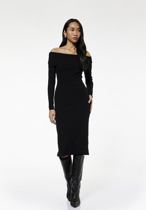 Vrouw draagt een nauwsluitende, zwarte midi-jurk met blote schouders en zwarte kniehoge laarzen, staand met één hand in de zak tegen een witte achtergrond.