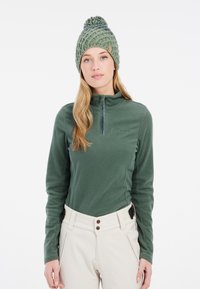 Pullover in pile verde scuro con zip a un quarto e tessuto testurizzato; abbinato a pantaloni beige chiaro e un berretto lavorato a maglia verde con pom-pom.