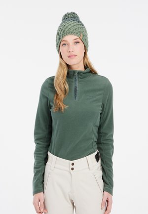 Dunkelgrüner Fleece-Pullover mit einem Viertelreißverschluss und strukturiertem Stoff; kombiniert mit hellbeigen Hosen und einer gestrickten grünen Mütze mit einem Bommel.