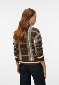 Brūns rakstainais džemperis ar baltām akcentiem, augstu apkakli un strukturētu adījumu; savienots ar tumšām džinsa biksēm.