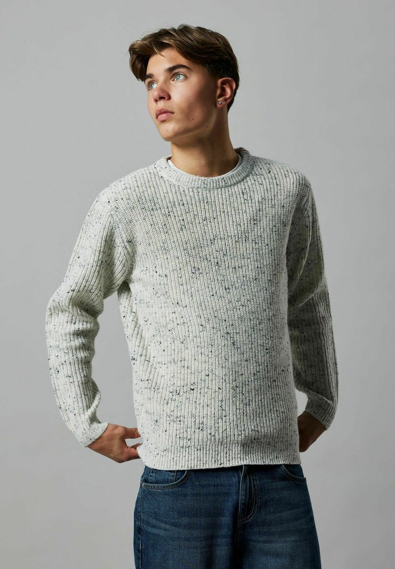 Helles grauer, gestrickter Pullover mit gerippter Textur und dezenten Sprenkeln, mit Rundhalsausschnitt und langen Ärmeln, kombiniert mit blauen Jeans.