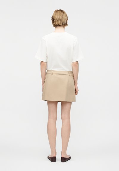 Person med kort blondt hår som har på seg en hvit kortermet topp, beige skjørt og mørke flate sko, stående med ryggen til på en hvit bakgrunn.