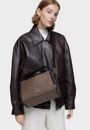 Mujer con chaqueta de cuero marrón oscuro y pantalones blancos, llevando un bolso Tous marrón con patrón y logo dorado al hombro.