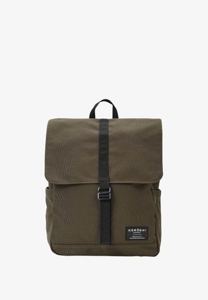 Mochila de lona verde oscura con tapa plegable, correa negra, bolsillos laterales y un parche de etiqueta. Cuenta con costuras duraderas y herrajes de metal.