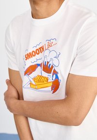 T-shirt bianca a maniche corte con un granchio fumetto che indossa un cappello da chef e tiene in mano un pezzo di formaggio con la scritta "NIKE", contornata da un bordo blu e arancione.