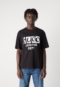 Black cotton t-shirt featuring a bold white graphic print of "HUGO CREATIVE DEPT." Short sleeves and a crew neckline. Casual fit.