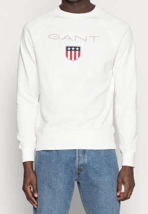 Mann in einem weißen GANT Sweatshirt mit einem rot-weiß-blauen Wappen-Logo, kombiniert mit blauen Jeans, stehend vor einem schlichten Hintergrund.