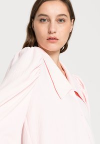 Blusa rosa-claro com decote de gola e mangas curtas bufantes. Feita em tecido texturizado com detalhes de botões na frente.