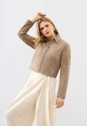 Femme portant une veste beige zippée ornée de clous et un pantalon blanc à jambes larges, levant une main au-dessus de sa tête devant un fond uni.