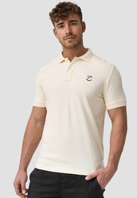 Helles beiges Polohemd aus weichem Strickstoff mit Kragen, kurzen Ärmeln und einem kleinen schwarzen Logo auf der Brust.