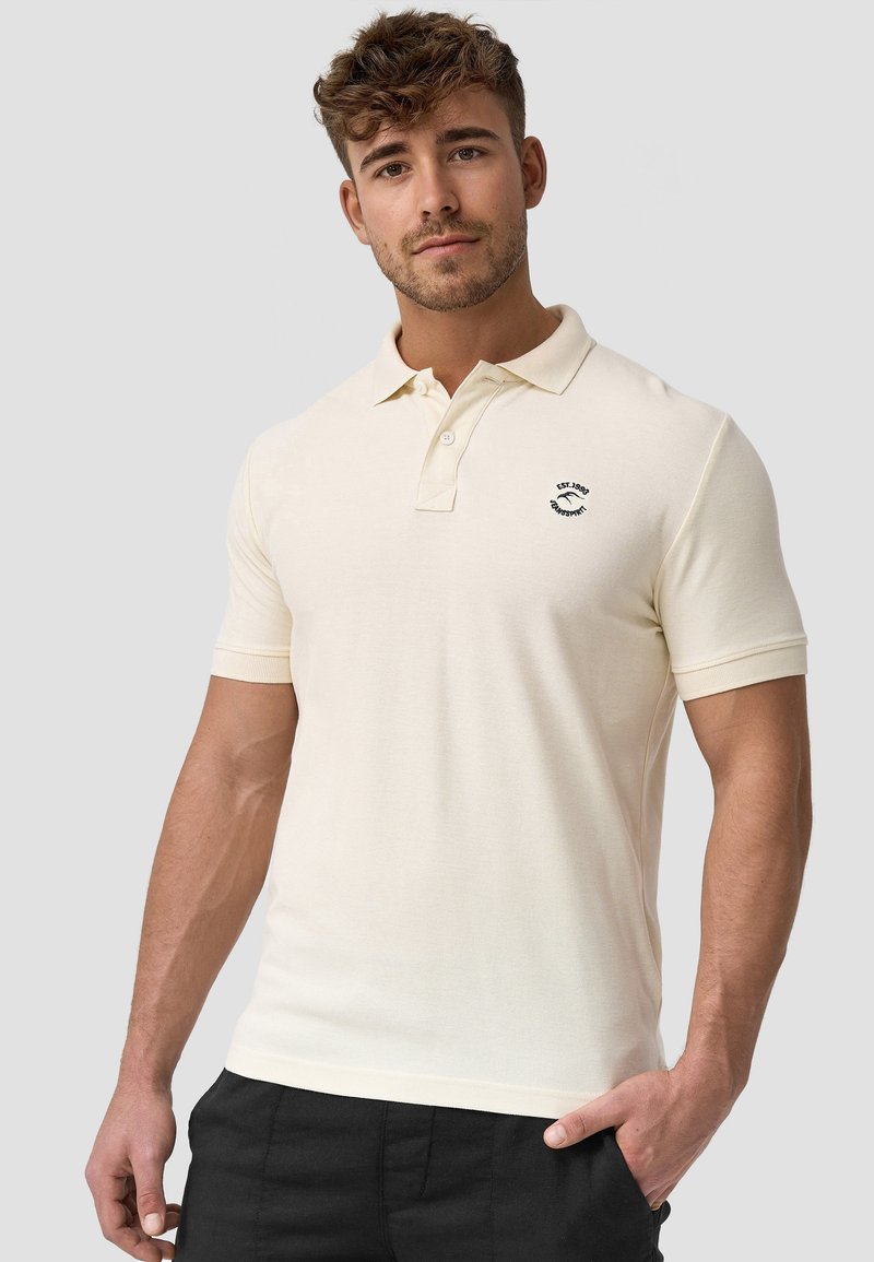 Helles beiges Polohemd aus weichem Strickstoff mit Kragen, kurzen Ärmeln und einem kleinen schwarzen Logo auf der Brust.