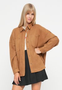 Brown faux suede shirt jacket with a collared neckline, two front pockets, and snap buttons, layered over a white top and black skirt.