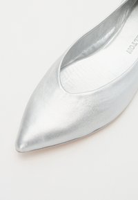 Chaussure plate en métal argenté avec un design à bout pointu. Texture lisse, style minimaliste et empeigne basse pour une apparence élégante.