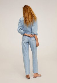 Žena s dlouhými vlnitými rudými vlasy, oblečená v světle modré džínové bundě a džínách, stojící bosá na jednobarevném béžovém pozadí, zády k fotoaparátu.