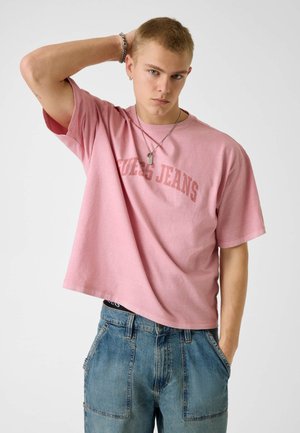 Giovane uomo con capelli corti biondi che indossa una T-shirt rosa "Guess Jeans", jeans di denim blu, collane d'argento e un braccialetto, posando con una mano dietro la testa.