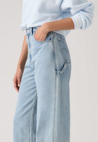 Άτομο που φοράει ανοιχτό μπλε φαρδιά τζιν παντελόνια με το χέρι στην μπροστινή τσέπη και ανοιχτό μπλε μακρυμάνικη μπλούζα μ προστά σε μονόχρωμο φόντο.