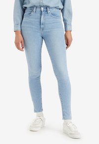 Jeans skinny de tiro alto en azul claro, hechos de mezclilla, con una textura suave y un diseño clásico de cinco bolsillos. Combinados con zapatillas blancas.