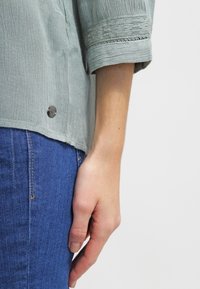 Blusa verde chiaro texturizzata con vestibilità rilassata, caratterizzata da maniche arrotolate e un bottone decorativo all'orlo. Abbinata a jeans in denim blu.